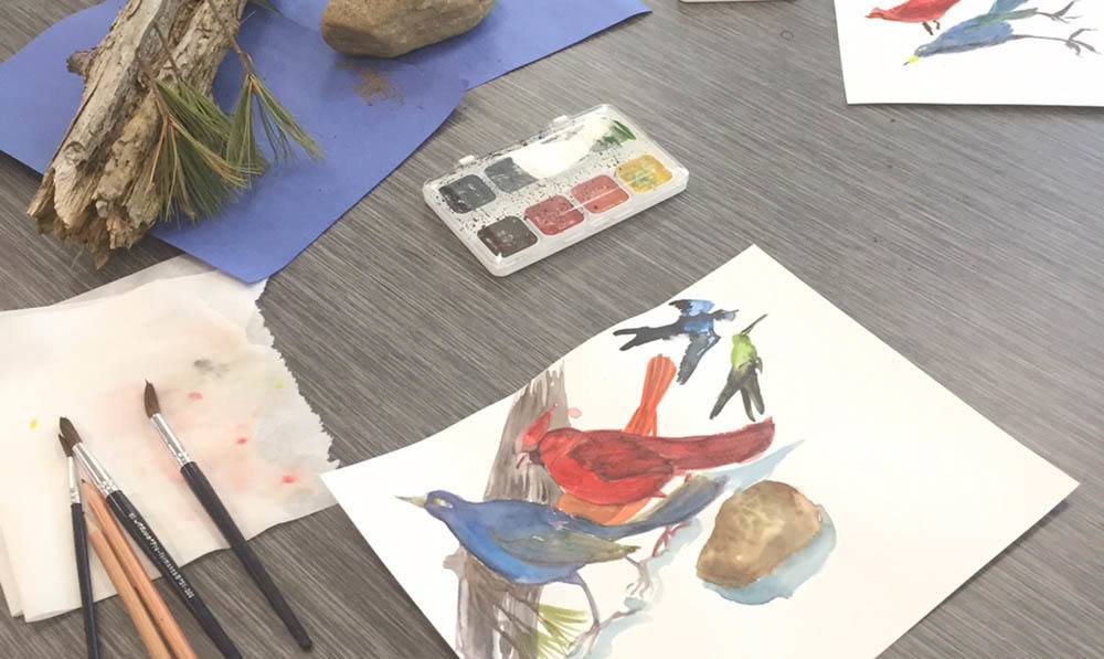 Paint palete, a paintbrush, and a painting of birds sits on a table in the Art Studio at The Ambassador